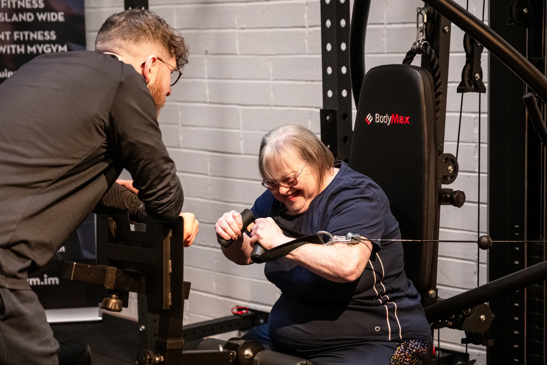 A woman with Down's syndrome smiling while using a BodyMax cable resistance machine assisted by a personal trainer in a gym, with a MyGym Isle of Man banner visible in the background